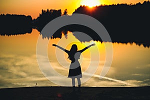 Silhouette of a young beautiful girl with hands up against the background of the sunset in the reflection of the pond