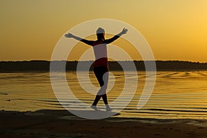 Silhouette of a young beautiful girl with hands up against the background of the sunset in the reflection of the sea coast