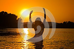 Silhouette of young beautiful woman in the river over sunset sky. Female perfect body contour at beach in twilight scenery