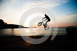 Silhouette of young caucasian biker jumping on sea coast in city on sunset sky background