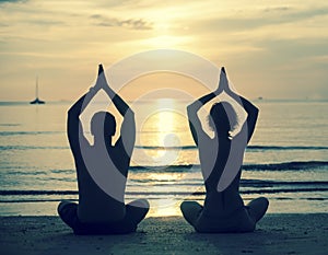 Silhouette of young couple practicing yoga on sea beach during sunset.