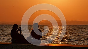 Silhouette of a young couple taking a selfie at sunset, with a sea and sunset in the background.