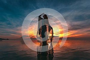 Silhouette of young fashionable woman standing in water on the beach at sunset