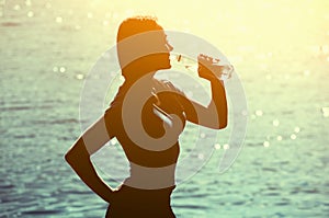 Silhouette of a young female athlete in tracksuit drinking water from a bottle on the beach in summer,