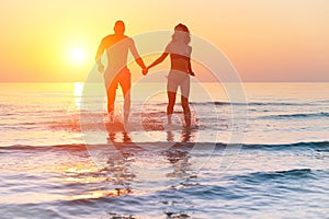 Silhouette of young fitness couple walking inside the water at sunrise - Multi race people having fun on vacation - Romantic and