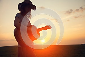 Silhouette of young free woman in straw hat playing country music on a guitar at sunset