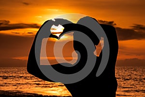 Silhouette of young girl with sunset on the beach, forming the shape of a heart with hands.