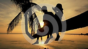 Silhouette of young happy father with his daughter sit on a palm tree during amazing golden sunset. Having fun on summer