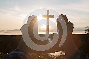 Silhouette of young human hands praying with a cross at sunrise, Christian Religion concept background