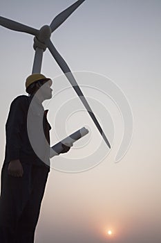 Silhouette of young male engineer holding a blueprint and standing beside a wind turbine at sunset