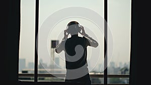 Silhouette of young man dancing ad listening music in wireles headphones stand on hotel room balcony