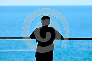 Silhouette of young man leaned on transparent glass handrail on above blue sparkling sea background