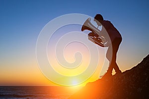 Silhouette of a young man playing the trumpet on rocky sea coast during sunset.