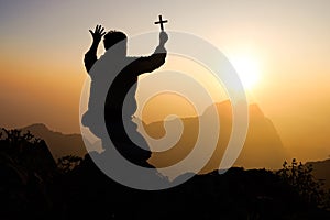 Silhouette of Young man praying and holding christian cross for worshipping God at sunset background. Christian Religion concept