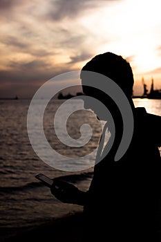 Silhouette of a young man in a sporty sweatshirt with a hood with a phone in hands against a beautiful sunset sky