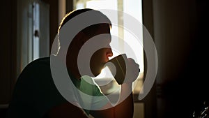 Silhouette of a young man in the sunbeams of dawn drinks hot coffee while sitting at home, camera movement