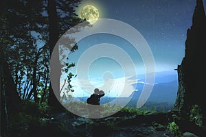 Silhouette of a young man who sits on the edge of the observation deck and enjoying night landscape with starry sky and full moon