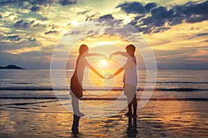 Silhouette of young romantic couple during tropical vacation, holding hands in heart shape on the ocean beach during sunset.