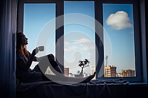 Silhouette of young slender girl sitting on the windowsill at home, side view, copy space. Outside the window blue sky and tall