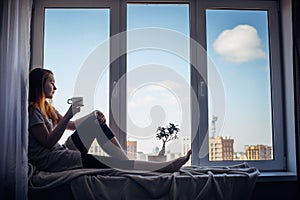 Silhouette of young slender girl sitting on the windowsill at home, side view, copy space. Outside the window blue sky and tall