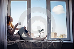 Silhouette of young slender girl sitting on the windowsill at home, side view, copy space. Outside the window blue sky and tall