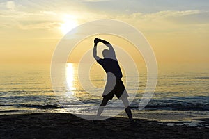 Silhouette of young sport man stretching leg after running workout outdoors on beach at sunset