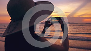 Silhouette of young tourist woman in hat taking photo with cellphone during sunset in ocean beach