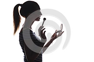 Silhouette of a young woman doing makeup in front of a pocket mirror, a girl with a brush and powder preens her face on a white