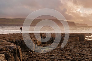 Silhouette of a young woman in Doolin, County Clare, Ireland, with Cliffs at background. Cloudy sky at sunset. Big rocks at
