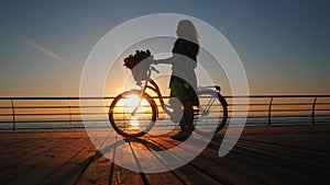 Silhouette of young woman in dress with vintage bicycle and bouquet of flowers walking on wooden embankment near sea