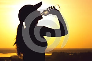 Silhouette of a woman drinking water after physical exercise in nature, sports female profile at sunset, concept of sport and rela