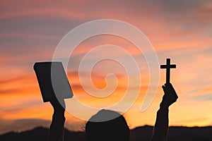 Silhouette of Young woman hands holding holy Bible and lift of christian cross, religion symbol in light and landscape over a