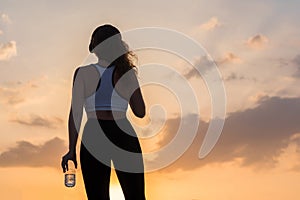 Silhouette young woman in the headphones in the sport clothes with bottle of clear mineral water on the sunset sky backgrounds.