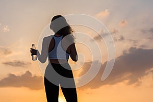 Silhouette young woman in the headphones in the sport clothes with bottle of clear mineral water on the sunset sky backgrounds.
