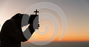 Silhouette of young woman kneeling down praying and holding christian cross for worshipping God at sunset background. concept of