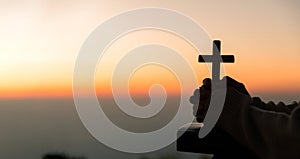 Silhouette of young woman kneeling down praying and holding christian cross for worshipping God at sunset background. concept of