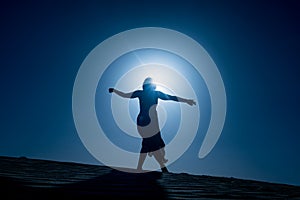 Silhouette of young woman with long skirt dancing in evocative and confident way on top of desert dune at night