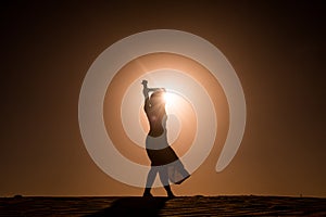 Silhouette of young woman with long skirt dancing in evocative and confident way on top of desert dune at sunset with sun high in