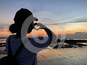 Silhouette of a young woman making heart love sign against colorful sunset on the beach. Showing love and kindness concept.