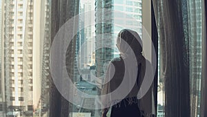 Silhouette of young woman opens curtains on the big window stretches arms and looking out her apartment on the city