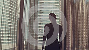 Silhouette of young woman opens curtains on the big window stretches arms and looking out her apartment on the city