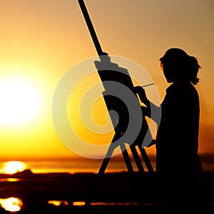 Silhouette of a young woman painting a picture on an easel on nature, girl figure with brush and artist`s palette engaged in art