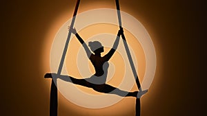 Silhouette of a young woman performs acrobatic elements and twine on airy silk. An aerial gymnast demonstrates