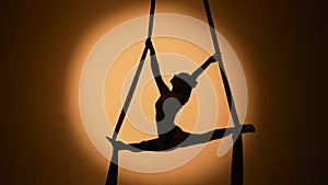 Silhouette of a young woman performs acrobatic elements and twine on airy silk. An aerial gymnast demonstrates