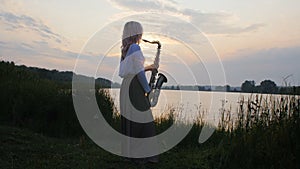 Silhouette of a young woman playing on the saxophone near the riverbank at dawn, a beautiful girl in a long dress with a wind musi