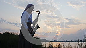 Silhouette of a young woman playing on the saxophone near the riverbank at dawn, a beautiful girl in a long dress with a wind musi