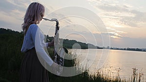 Silhouette of a young woman playing on the saxophone near the riverbank at dawn, a beautiful girl in a long dress with a wind musi