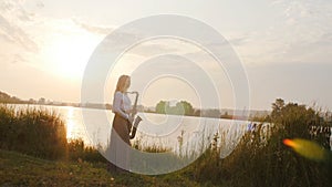 Silhouette of a young woman playing on the saxophone near the riverbank at dawn, a beautiful girl in a long dress with a wind musi