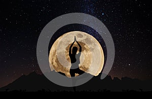 Silhouette young woman practicing yoga pose at night under full moon with sky full of stars