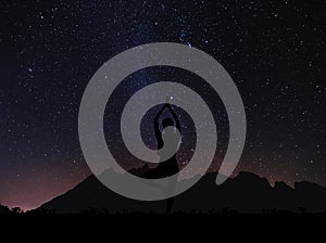 Silhouette young woman practicing yoga pose at night under sky full of stars with colorful milky way